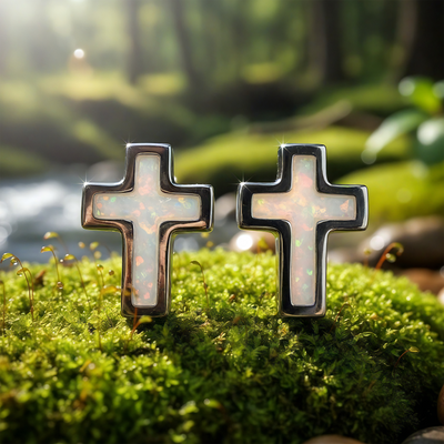 A pair of cross-shaped stud earrings with shimmering white opal inlay in sterling silver, standing on moss
