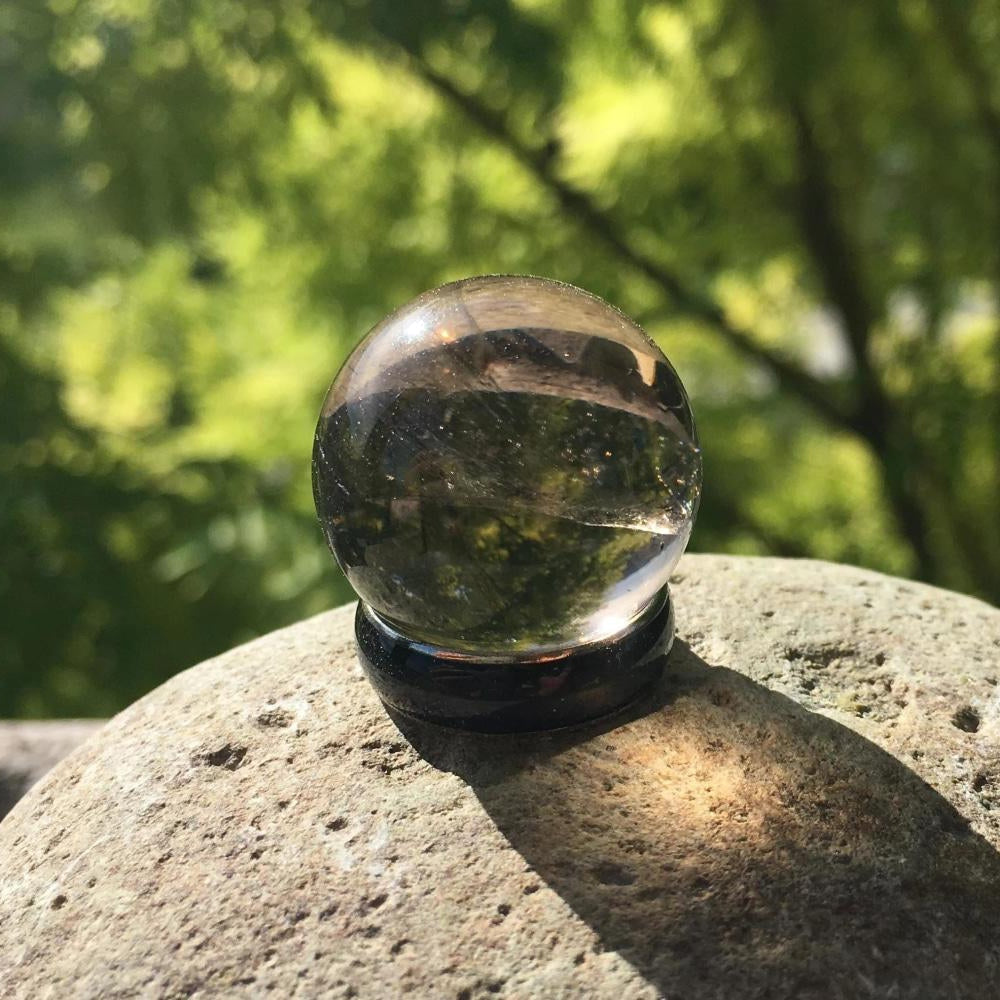 Polished translucent smoky quartz sphere on a black stand, resting on a stone in sunlight