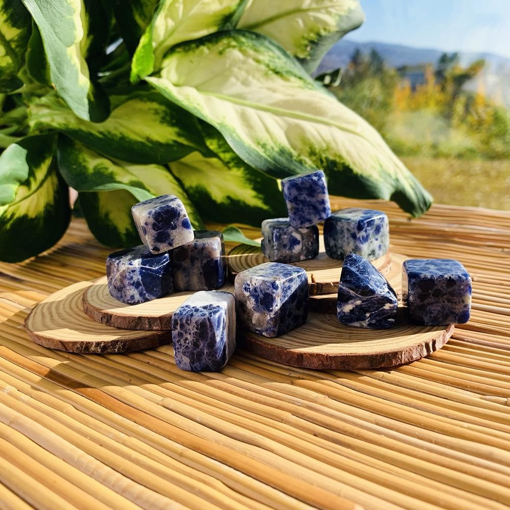 Royal blue sodalite tumbled cubes veined with white, stacked on wood slices on a bamboo mat beside a leafy plant