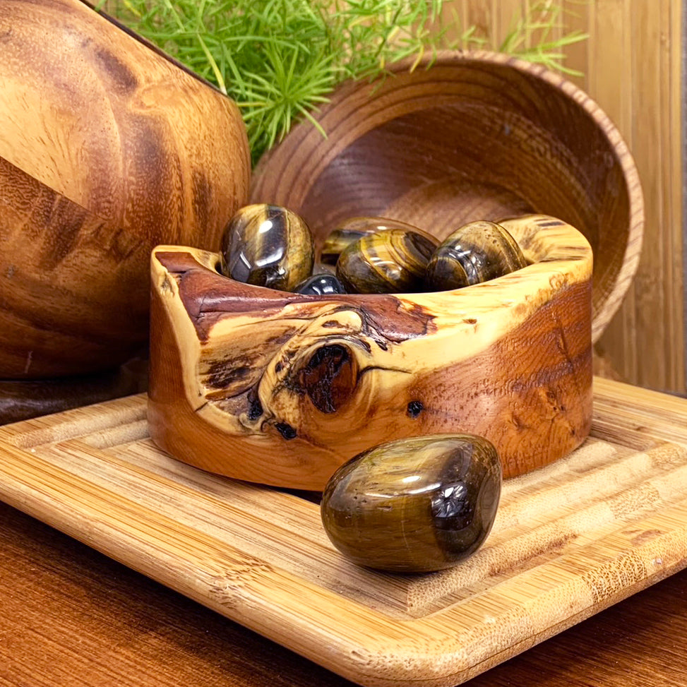 A tumbled tiger’s eye ellipse with golden-brown bands resting in front of a wooden bowl of more tiger’s eye stones