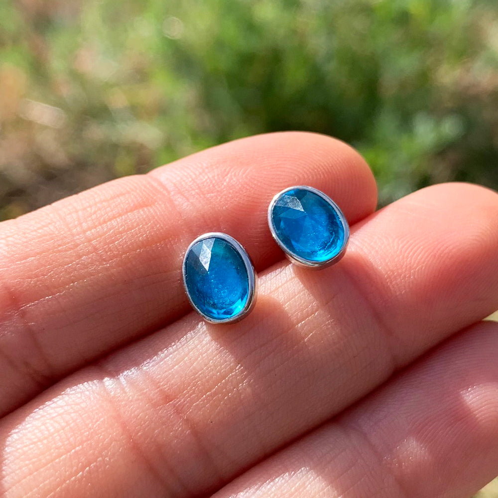 Two oval faceted blue topaz studs in sterling silver bezels resting on fingertips in sunlight